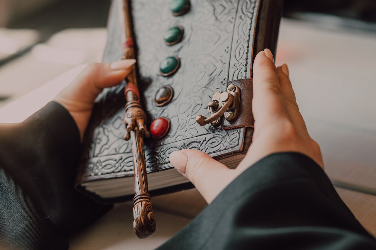 Close-up of hands holding an embossed leather book with a wand and gemstones.